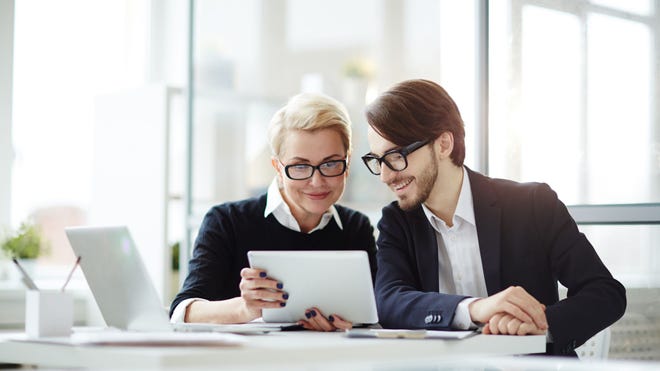 A man and woman in an office setting look smiling at the screen of a tablet computer.